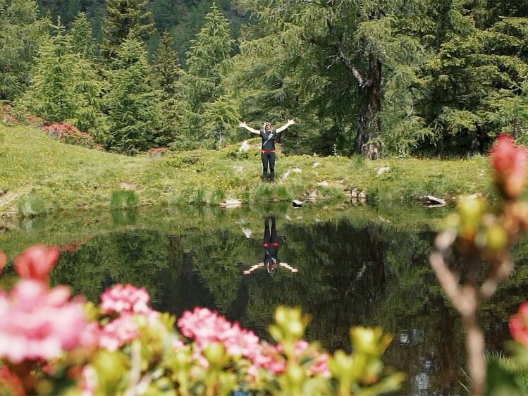 Person standing with arms raised, reflected in still lake surrounded by lush, green forest and colorful flowers.