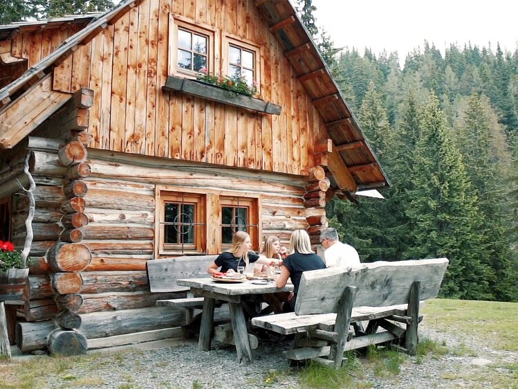 A rustic wooden cabin with a group of people dining outdoors on a grassy field surrounded by a forested landscape. | © Urlaub am Bauernhof Kärnten