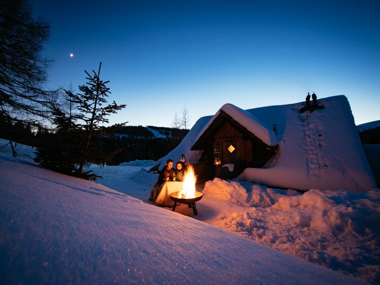 A cozy, snow-covered cabin with a warm fireplace, surrounded by a serene winter landscape under a starry sky. | © Urlaub am Bauernhof Kärnten / Daniel Gollner