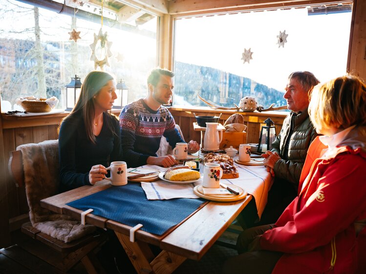 Cozy wooden cabin interior, group of people enjoying winter snacks and drinks, scenic mountain landscape visible through window. | © Urlaub am Bauernhof Kärnten / Daniel Gollner