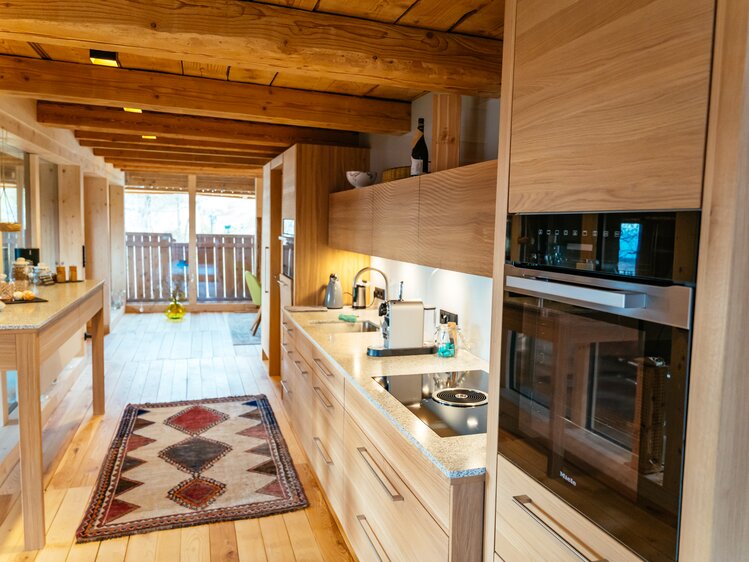 Rustic wooden kitchen with modern appliances, open floor plan, and colorful patterned rug. Glass doors lead to a balcony or deck. | © Urlaub am Bauernhof Kärnten / Daniel Gollner