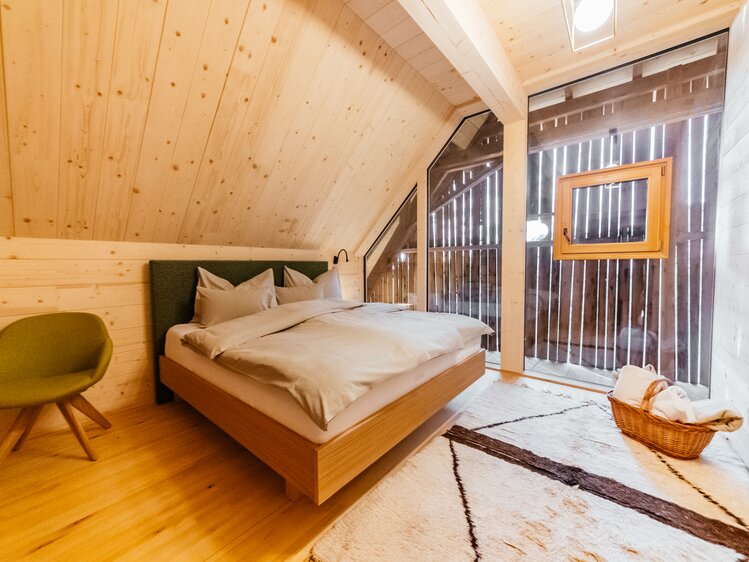 Cozy wooden bedroom with large bed, green armchair, and woven rug. Exposed beams and windows provide natural light. | © Urlaub am Bauernhof Kärnten / Daniel Gollner