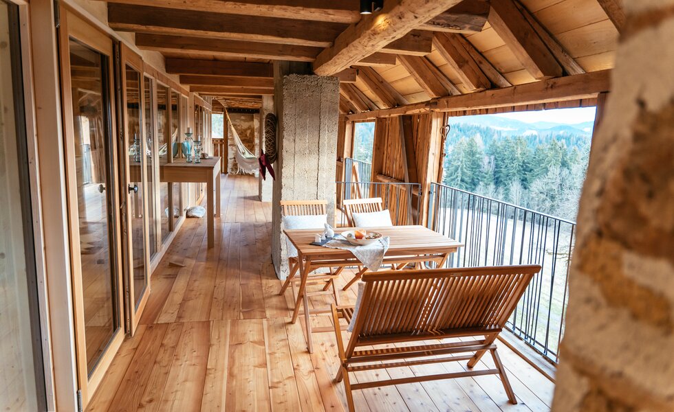 Wooden mountain cabin with a balcony overlooking a forested landscape, featuring a dining table and chairs. | © Urlaub am Bauernhof Kärnten / Daniel Gollner