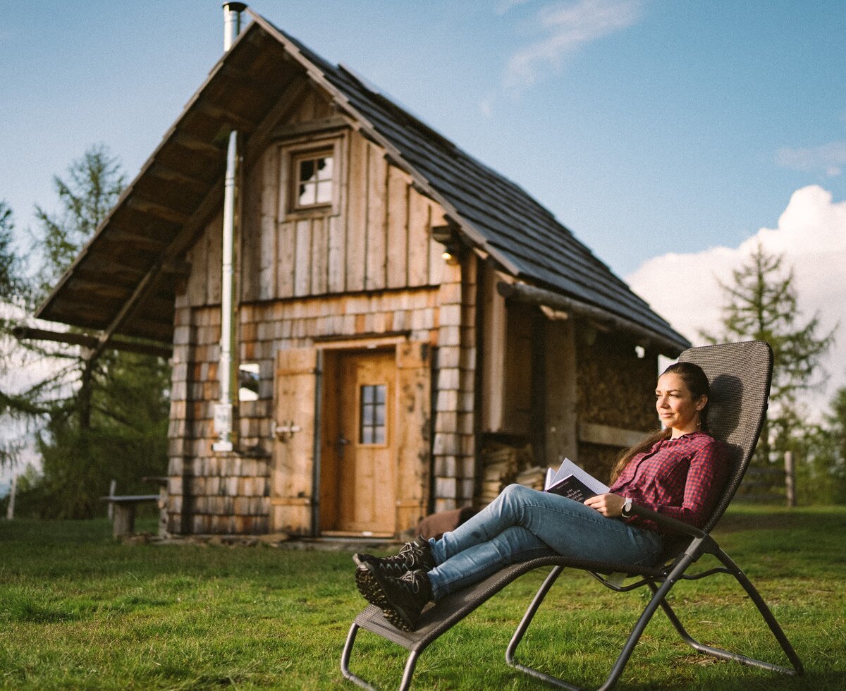 Rustic mountain cabin, woman relaxing in chair reading book, grassy field, evergreen trees, and cloudy sky backdrop. | © Urlaub am Bauernhof / Daniel Gollner