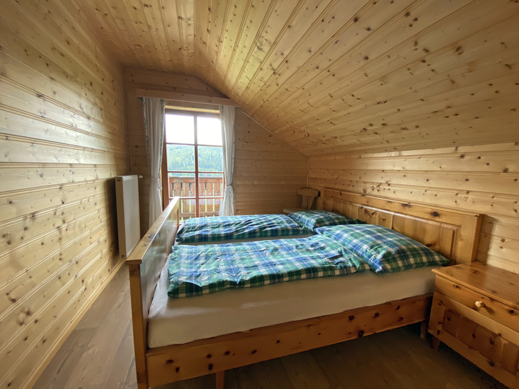 Cozy wooden chalet bedroom with plaid bedding, slanted ceiling, and large window overlooking the scenery. | © Jasmin Kreulitsch
