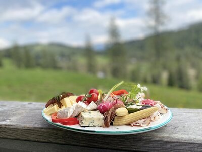 A plate filled with various fresh vegetables, cheeses, and charcuterie, set against a scenic mountain landscape. | © Urlaub am Bauernhof Kärnten / Jasmin Kreulitsch