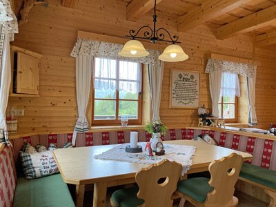 Cozy wooden cabin interior with a dining table, benches, decorative curtains, and a pendant light fixture. | © Urlaub am Bauernhof Kärnten / Jasmin Kreulitsch