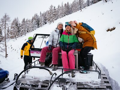 Group of people in winter outfits taking a selfie on a snow-covered mountain path, seated on a snow vehicle. | © Urlaub am Bauernhof Kärnten/ Daniel Gollner