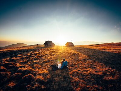 Ein romantischer Sonnenuntergang auf der Alm in Kärnten | © Urlaub am Bauernhof Kärnten/ Daniel Gollner Scenic sunset over rugged countryside with two small wooden cabins and a couple sitting in the foreground. | © Urlaub am Bauernhof Kärnten/ Daniel Gollner