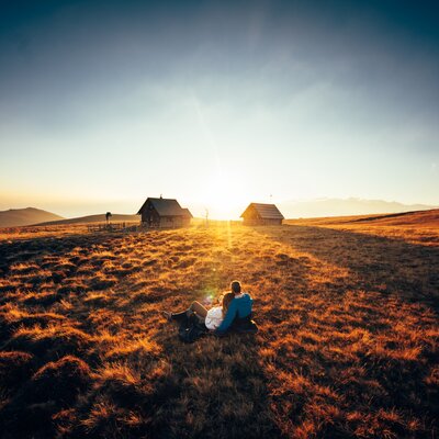 Scenic sunset over rugged countryside with two small wooden cabins and a couple sitting in the foreground. | © Urlaub am Bauernhof Kärnten/ Daniel Gollner 