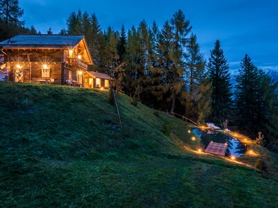 Rustic log cabin surrounded by tall pines, with a path leading to a pond and lights illuminating the tranquil scene. | © Günter Standl