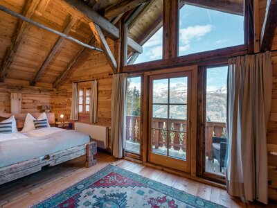 Cozy wooden cabin interior with a large bed, patterned rug, and panoramic mountain views through the windows. | © Günter Standl 