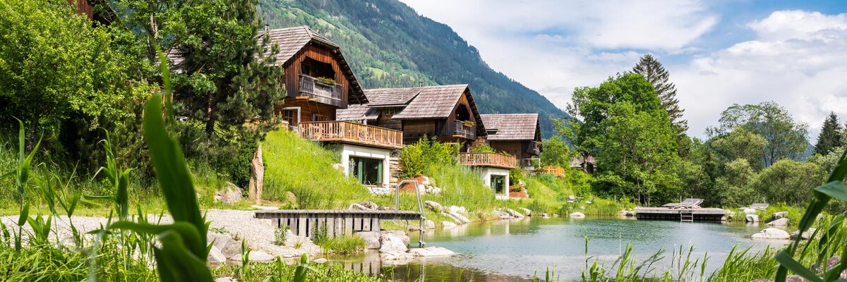 Picturesque wooden chalets nestled by a tranquil pond, surrounded by lush greenery and majestic mountains under a blue sky. | © Michael Stabentheiner