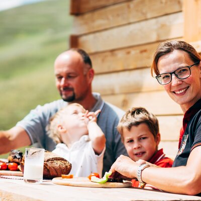 Happy family enjoying a summer meal outdoors in a rustic setting, with young children and adults relaxing together. | © Urlaub am Bauernhof Kärnten / Daniel Gollner