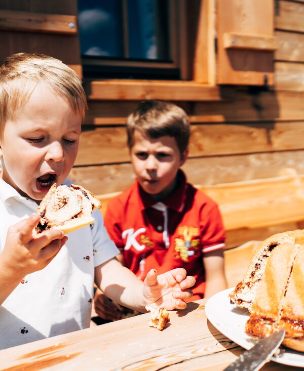 Two young boys enjoying sweet pastries outdoors, surrounded by rustic wooden structures. | © Urlaub am Bauernhof Kärnten / Daniel Gollner