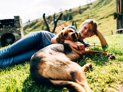 Cheerful woman cuddles with friendly dog on idyllic grassy slope near rustic wooden structures. | © Urlaub am Bauernhof Kärnten / Daniel Gollner