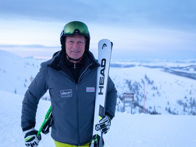 Person wearing ski attire, holding skis, standing on snowy mountain against cloudy sky. | © BRM / Mathias Prägant
