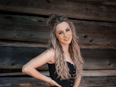 Woman with long gray hair wearing a black fringe top and pink metallic shorts, posing against a rustic wooden wall. | © Lambauer Entertainment / Christoph Hatheuer