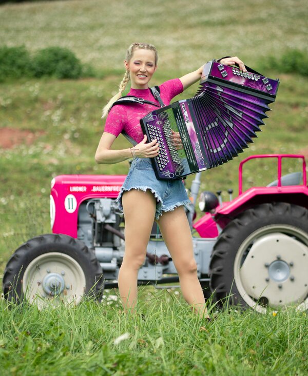 Smiling woman posing with colorful accordion on a red tractor in a grassy field. | © Lambauer Entertainment / Christoph Hatheuer