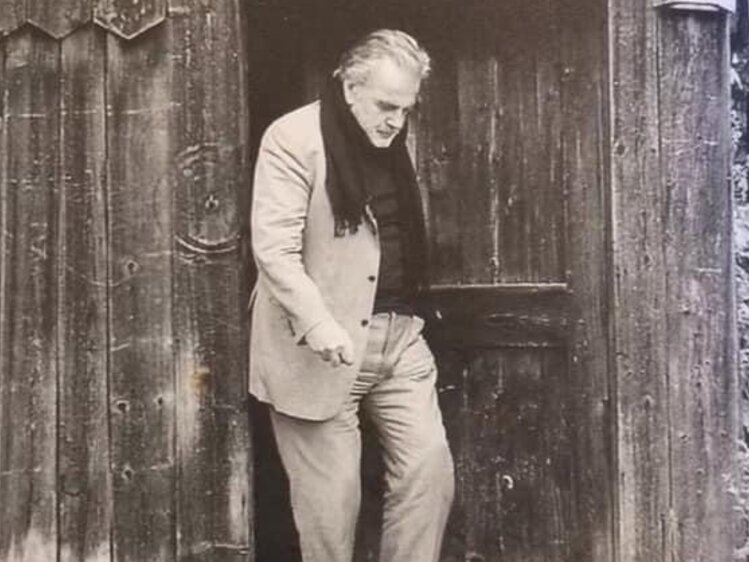 Elderly man in casual clothing standing in the doorway of a rustic wooden building, surrounded by decorative plants. | © Urlaub am Bauernhof / Schell