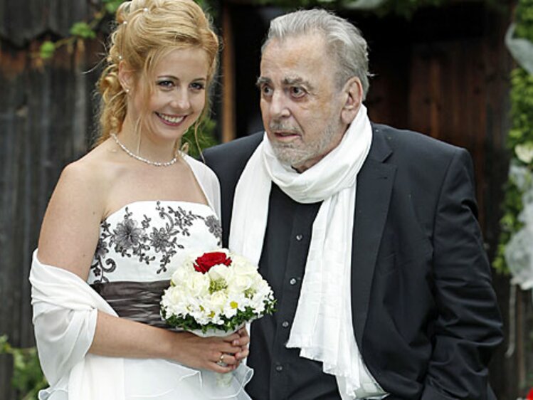Elderly gentleman in formal attire escorting a smiling young woman in a floral-patterned wedding dress, holding a bouquet of flowers. | © Urlaub am Bauernhof / Schell