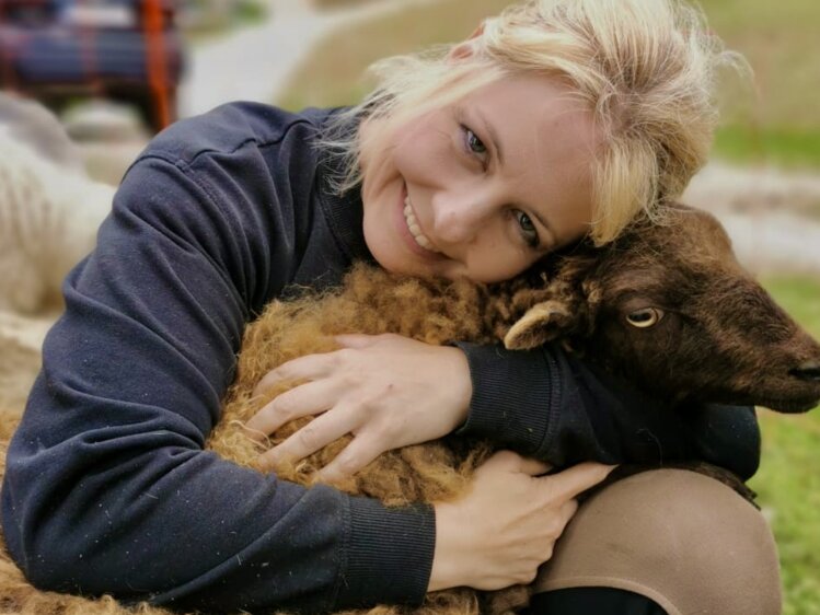Smiling woman embracing a fluffy sheep in a grassy, forested setting. | © Urlaub am Bauernhof / Schell