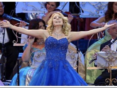 Sängerin Iva Schell | © Urlaub am Bauernhof / Schell Elegant woman in a sparkling blue gown performing on a stage with an orchestra in the background. | © Urlaub am Bauernhof / Schell