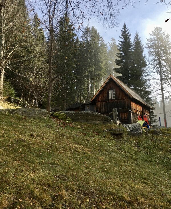 Rustic wooden cabin in a peaceful forest setting, with hikers exploring the surroundings on a misty day. | © Urlaub am Bauernhof / Schell
