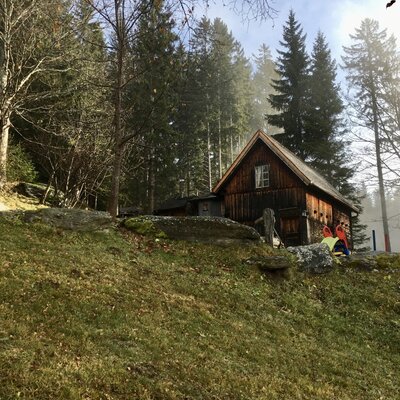 Rustic wooden cabin in a peaceful forest setting, with hikers exploring the surroundings on a misty day. | © Urlaub am Bauernhof / Schell