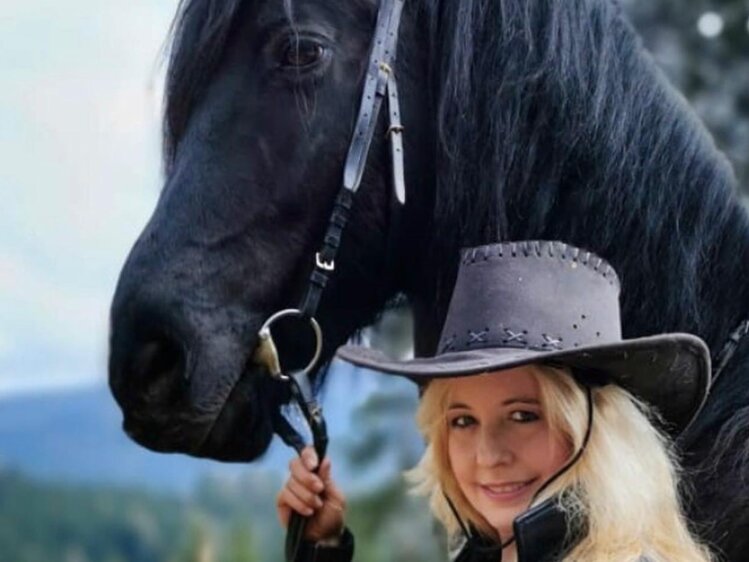 A woman in a cowboy hat stands next to a large black horse in a mountainous outdoor setting. | © Urlaub am Bauernhof / Schell