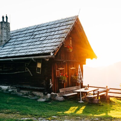 Rustic wooden cabin with slate roof, porch, and scenic backdrop of forest and mountain at sunset. | © Urlaub am Bauernhof Kärnten / Daniel Gollner