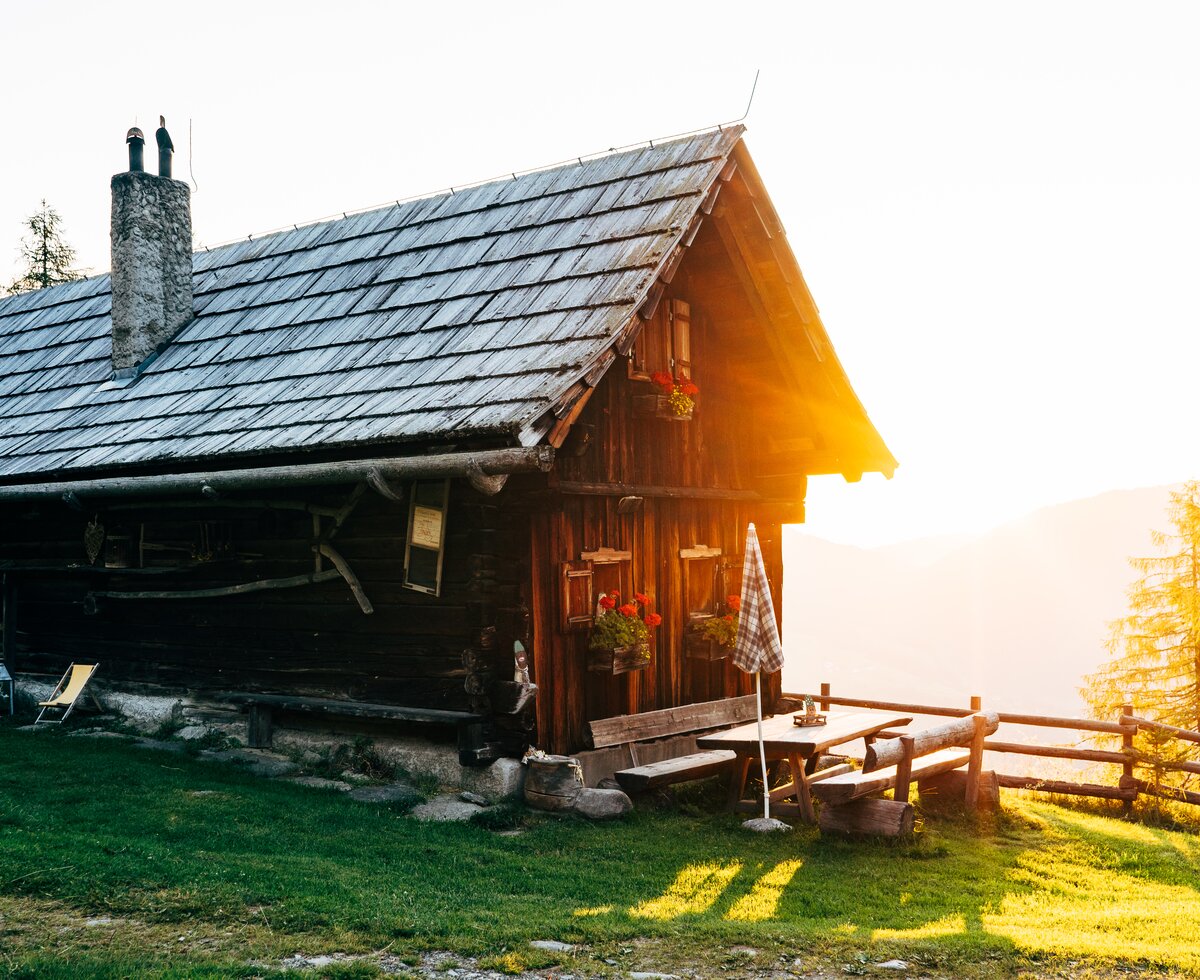 Rustic wooden cabin with slate roof, porch, and scenic backdrop of forest and mountain at sunset. | © Urlaub am Bauernhof Kärnten / Daniel Gollner