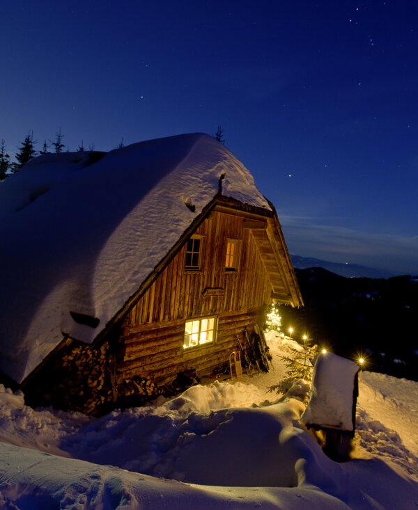 Cozy wooden cabin nestled in snowy landscape, illuminated at night under starry sky. Serene winter scene. | © Urlaub am Bauernhof/ Tom Lamm