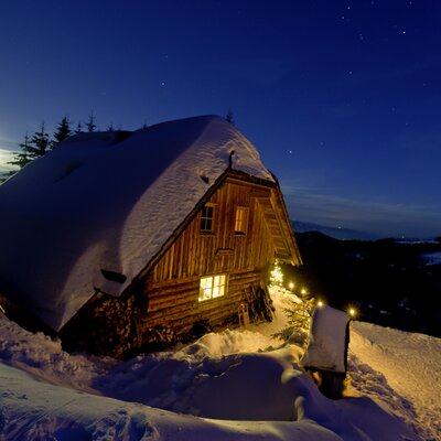 Cozy wooden cabin nestled in snowy landscape, illuminated at night under starry sky. Serene winter scene. | © Urlaub am Bauernhof/ Tom Lamm