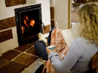 Frau wärmt sich am Ofen auf der Alm | © Urlaub am Bauernhof/ Tom Lamm Person sits by fireplace, reading book, cozy indoor scene with warm lighting. | © Urlaub am Bauernhof/ Tom Lamm