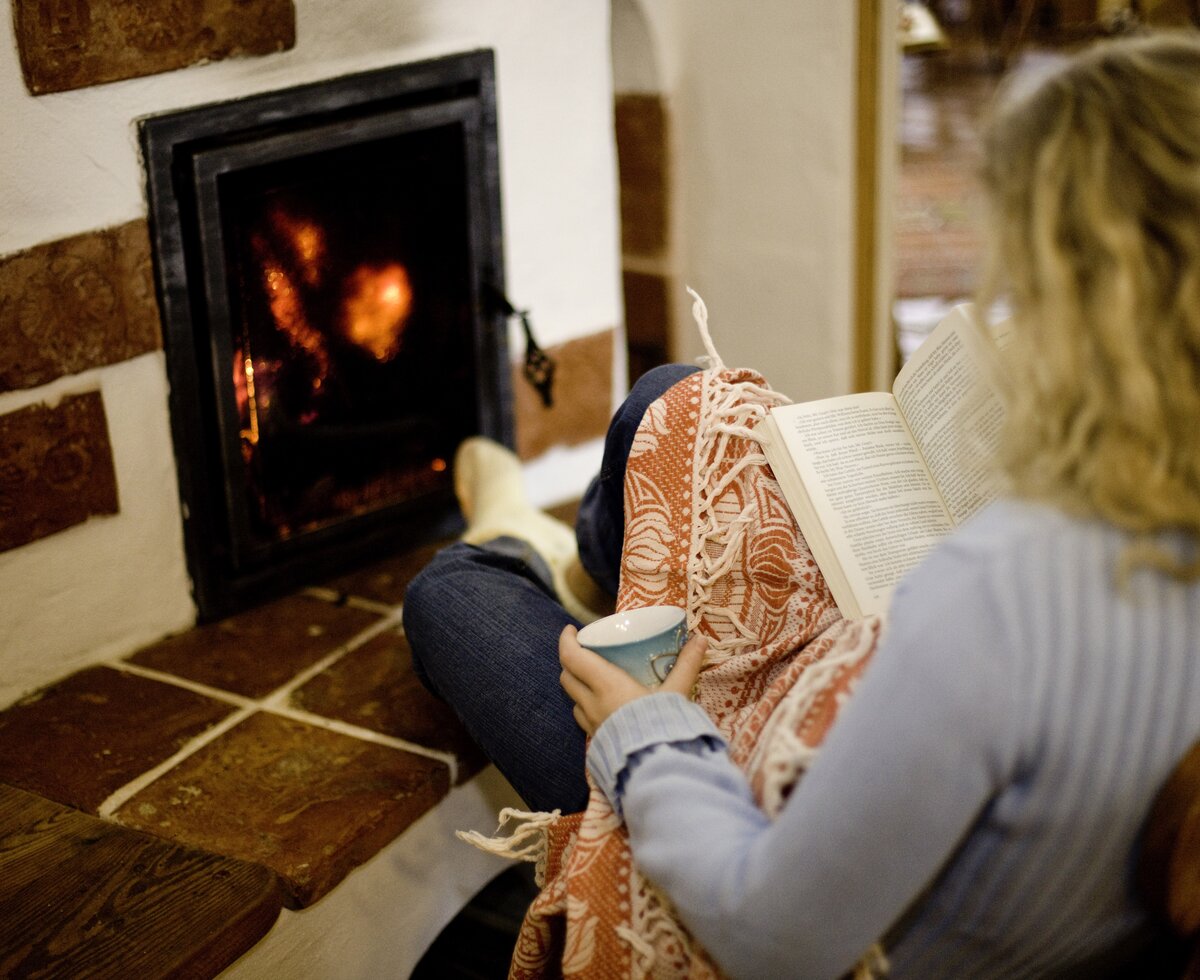 Person sits by fireplace, reading book, cozy indoor scene with warm lighting. | © Urlaub am Bauernhof/ Tom Lamm