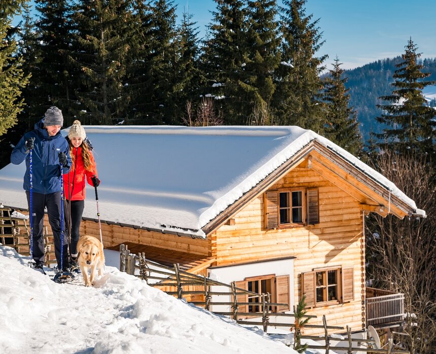 Snowy log cabin in forest with couple and dog hiking, warm winter scenery with trees and mountains in background. | © Michael Stabentheiner