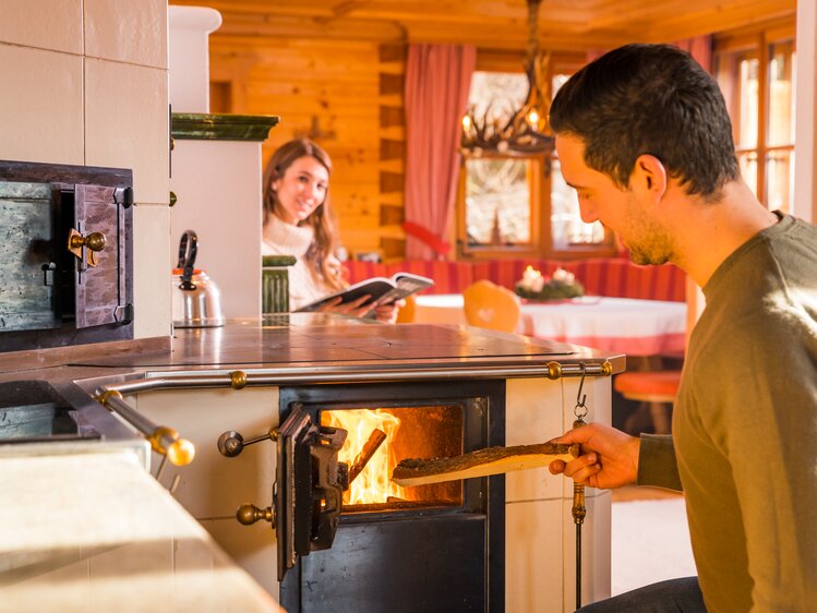 Couple in a cozy wooden cabin, man tending to fireplace while woman holds a digital device, warm atmosphere. | © Michael Stabentheiner
