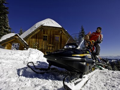 Rustic wooden huts, snow-covered trees, people on snowmobiles enjoying winter vacation in snowy landscape.
