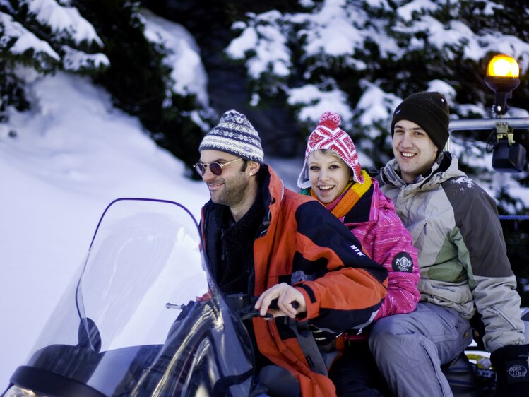 Three people in winter gear on a snowmobile surrounded by snowy trees at night. | © Urlaub am Bauernhof / Tom Lamm