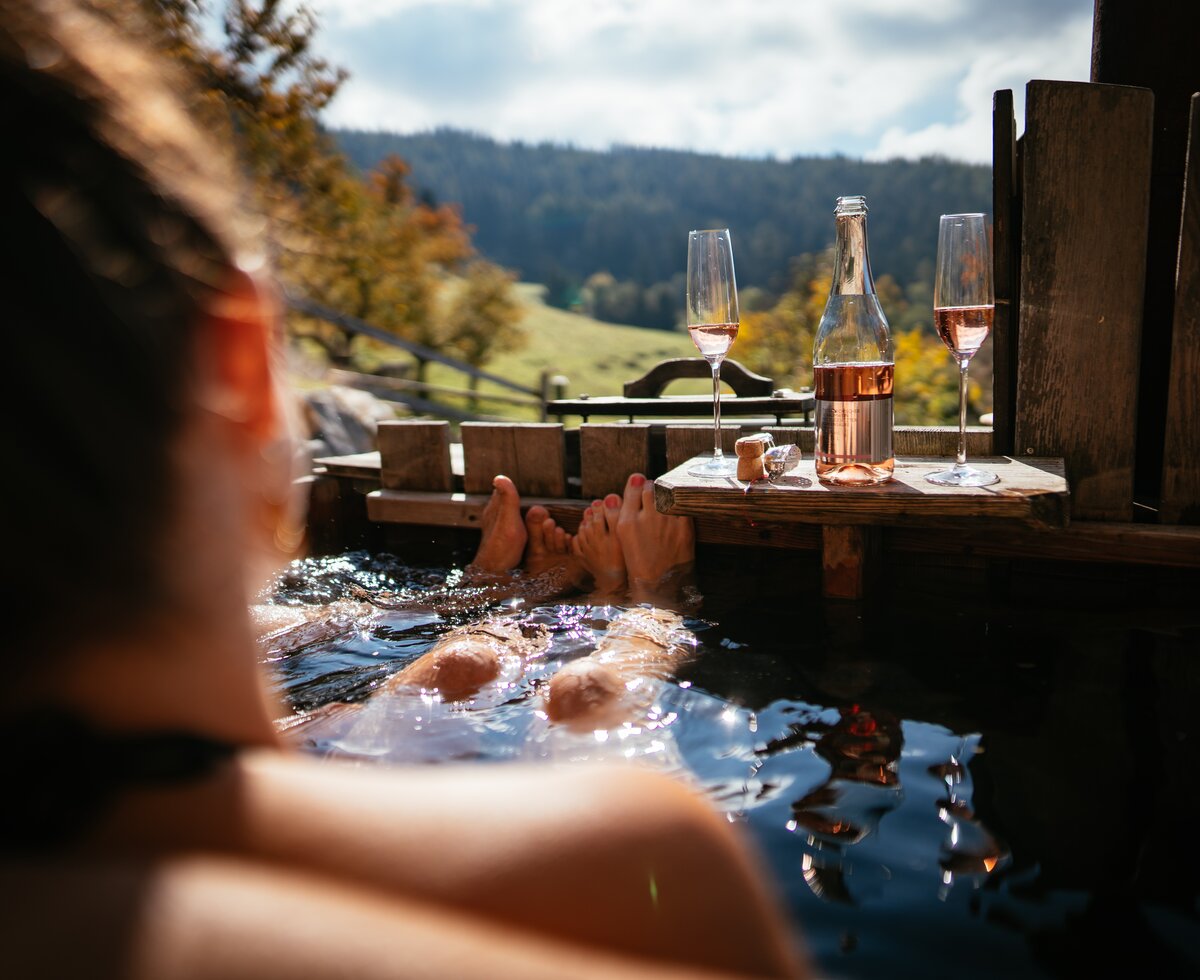 Outdoor hot tub with wine glasses, mountain landscape in background, feet visible in the water. | © Urlaub am Bauernhof / Daniel Gollner