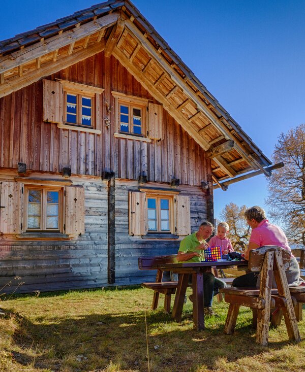 Rustic wooden cabin with family seated at table enjoying autumn landscape overlooking lake.