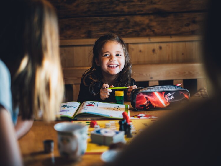 A young girl smiling happily while surrounded by various school supplies and toys in a rustic, wooden room. | © Urlaub am Bauernhof Salzburg / Daniel Gollner 