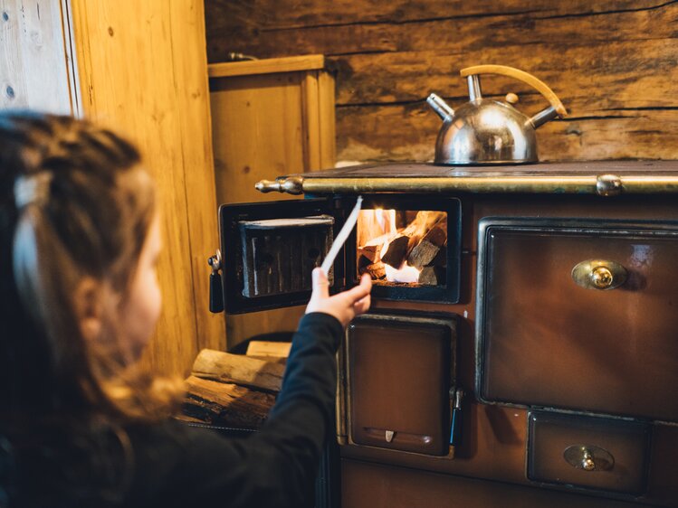 Rustically furnished room, wood-burning stove lit, kettle on top, person tending to the fire. | © Urlaub am Bauernhof Salzburg / Daniel Gollner 