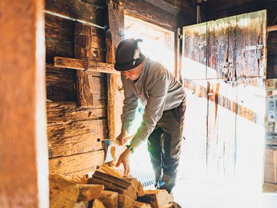 Man in workwear chopping firewood in a rustic wooden cabin interior, with shelves and art on the walls. | © Urlaub am Bauernhof Salzburg / Daniel Gollner 