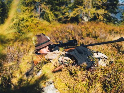 Jäger auf der Pirsch | © Urlaub am Bauernhof Salzburg / Daniel Gollner A person lying on the ground in camouflage clothing, aiming a rifle with a scope towards the distant forest landscape. | © Urlaub am Bauernhof Salzburg / Daniel Gollner