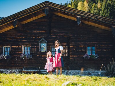 Mädchen im Dirndl vor der Almhütte | © Urlaub am Bauernhof Salzburg / Daniel Gollner Two women in traditional Bavarian clothing stand in front of a rustic wooden chalet with floral window boxes and a snowy mountain backdrop. | © Urlaub am Bauernhof Salzburg / Daniel Gollner