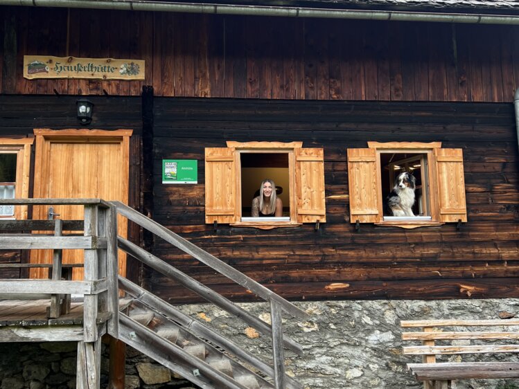 Weathered wooden wall with stacked wooden crates, a cat peering out, and a metal ladder on the side. | © Urlaub auf der Alm / Julia Winkler