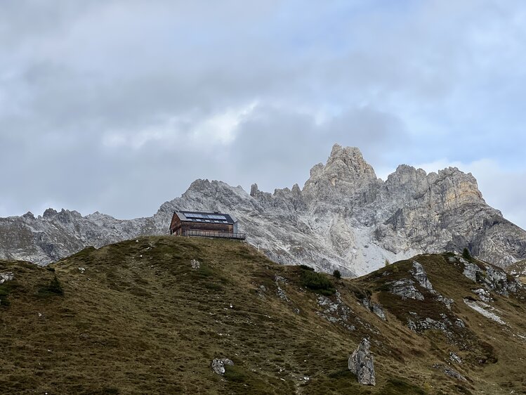 Wooden cabin on a grassy hill overlooking majestic snow-capped mountains under a cloudy sky. | © Urlaub auf der Alm / Julia Winkler