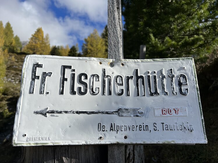 A wooden sign indicating the "Fr. Fischerhiitte" hiking trail, with a worn and weather-beaten appearance, set against a backdrop of autumn trees and a blue sky. | © Urlaub auf der Alm / Julia Winkler